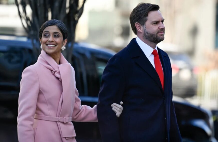 Usha Vance, Second Lady of the United States, smiling and gently cradling her baby bump, with a blurred Capitol and American flag background.